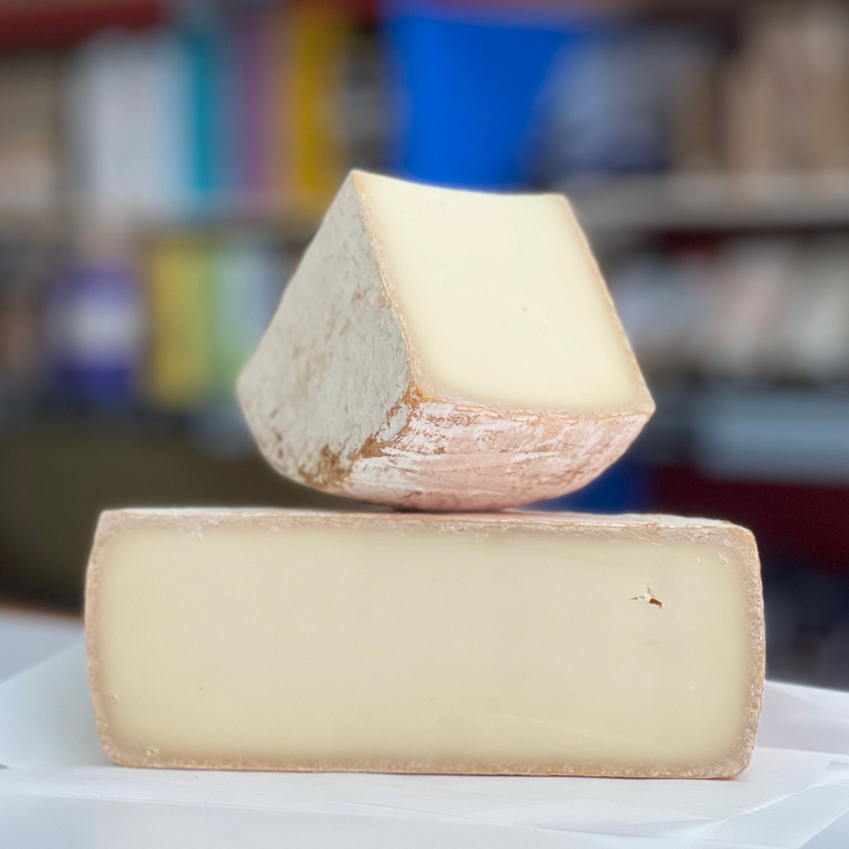 A sheep's milk cheese with an ochre rind cut in half and sitting on a counter. The background is blurred and a second piece of cheese is sitting on top pointing upward
