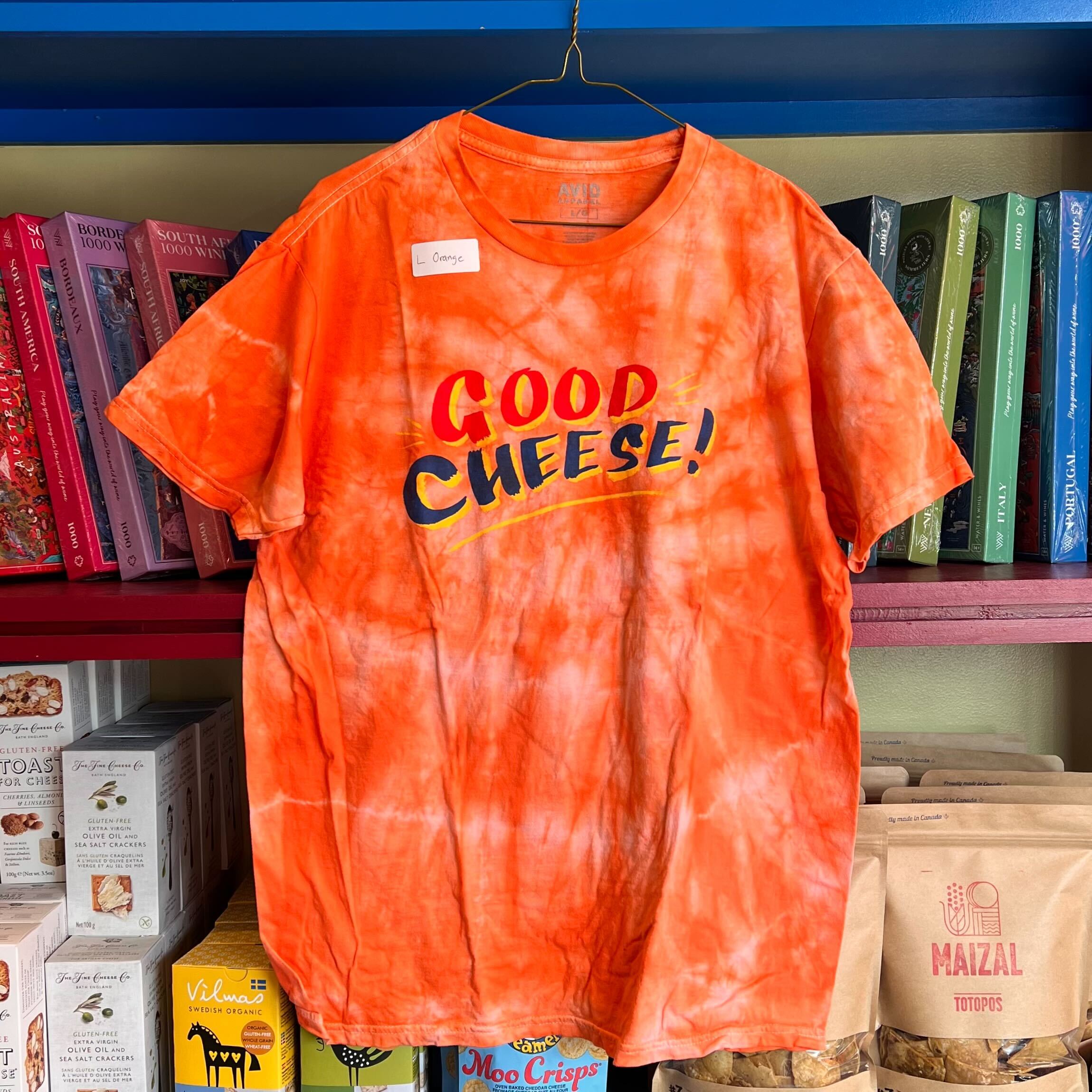 Orange tie-dye t-shirt with 'Good Cheese!' text on a shelf with books and snacks.