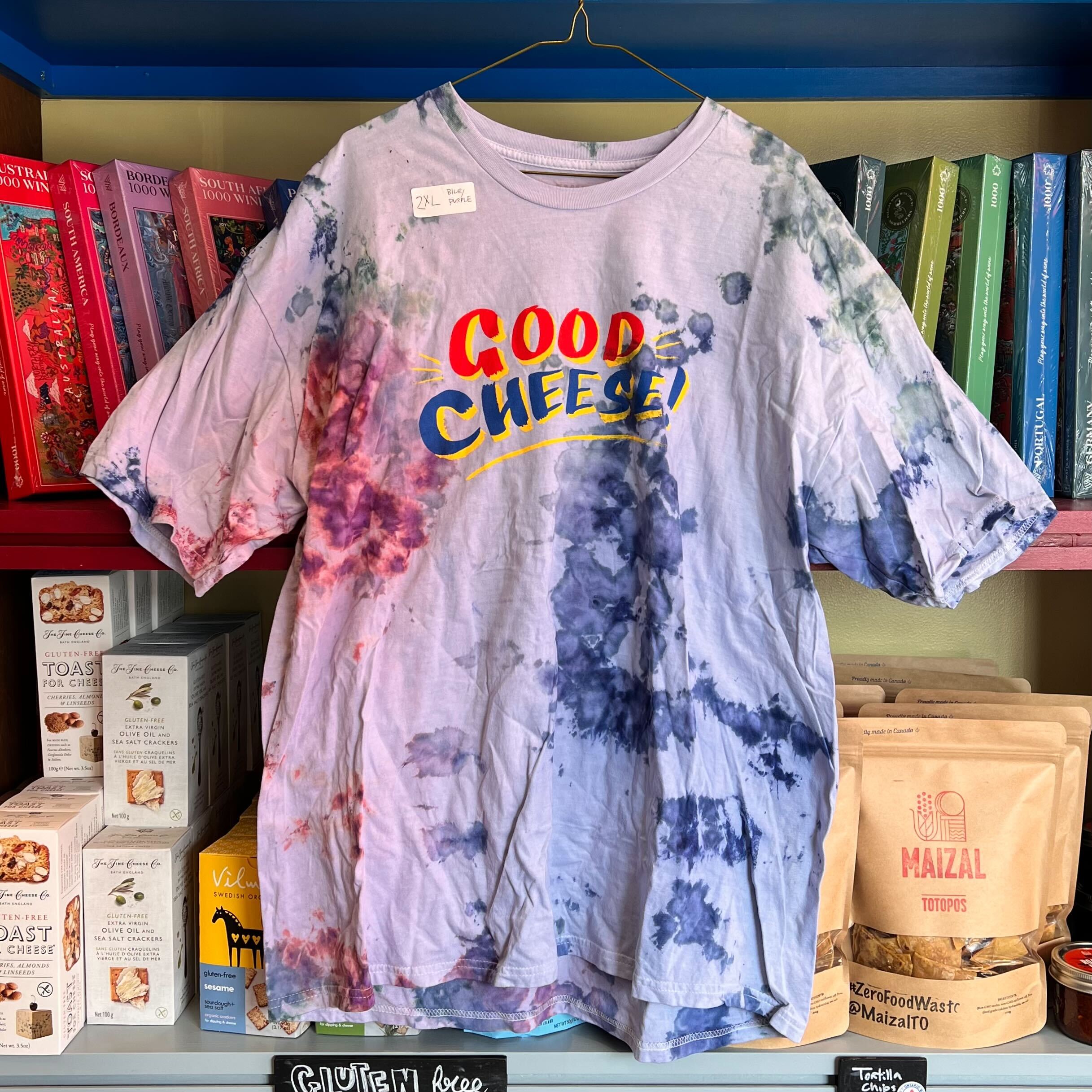 Tie-dye t-shirt with 'Good Cheese!' text hanging on a bookshelf with food products below.