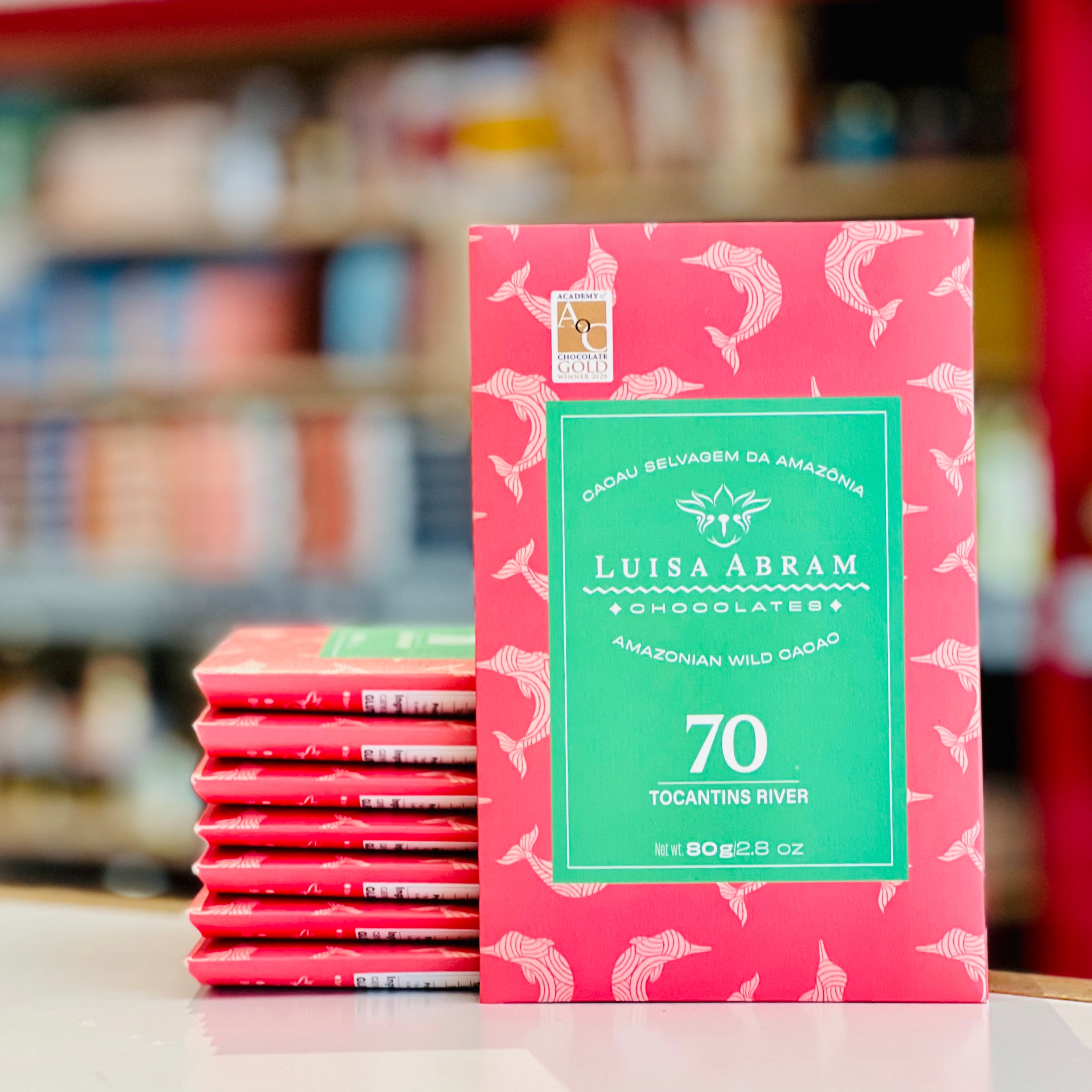 A red chocolate bar package labeled Luisa Abram Chocolates stands upright on a countertop, with several matching red bars stacked behind it. The package has a green central label and a pinkish-red background with white fish-like patterns.