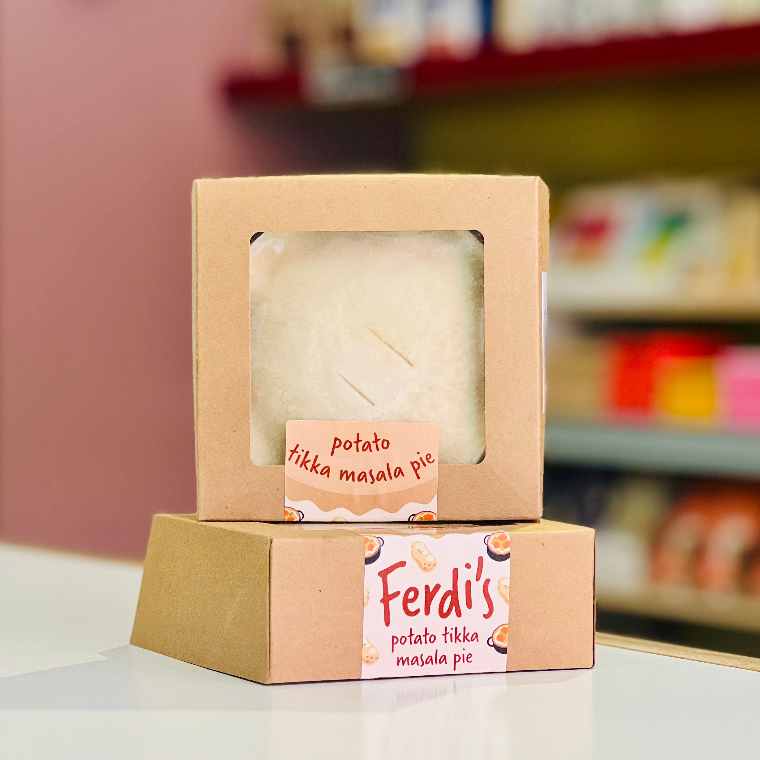 Two brown kraft paper boxes of Ferdi's Potato Tikka Masala Pie stacked on a white surface, with a blurred shop background.