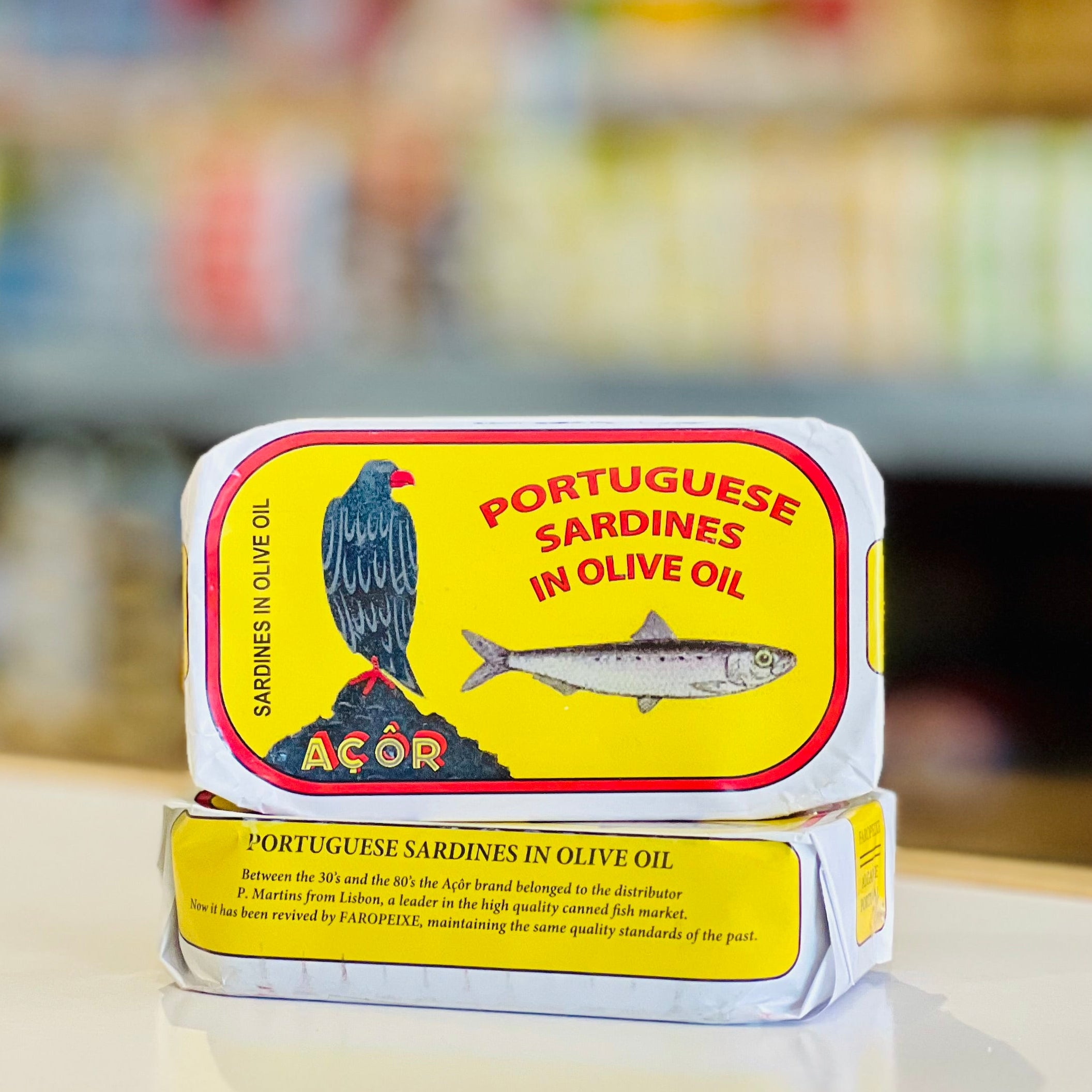 A tin of Portuguese Sardines in Olive Oil by Acor, stacked on another tin, set on a reflective white surface with a blurred supermarket background.