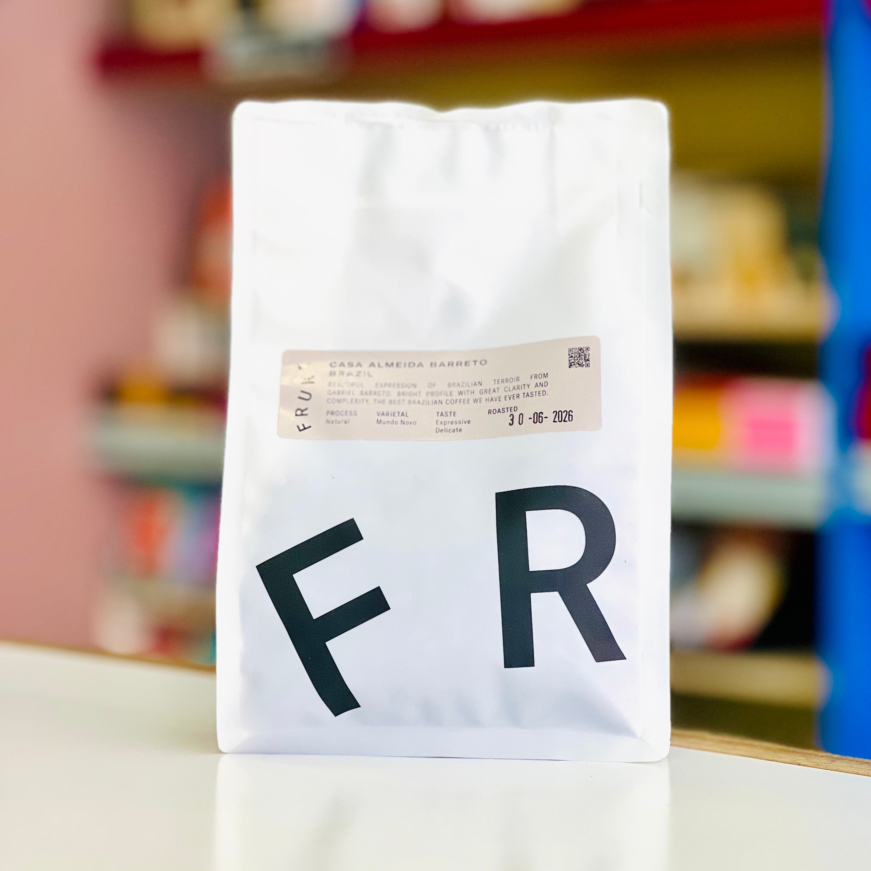 A white coffee bag with large black letters 'F R' on the front and a small beige label near the top, standing on a white surface with a blurred indoor background.