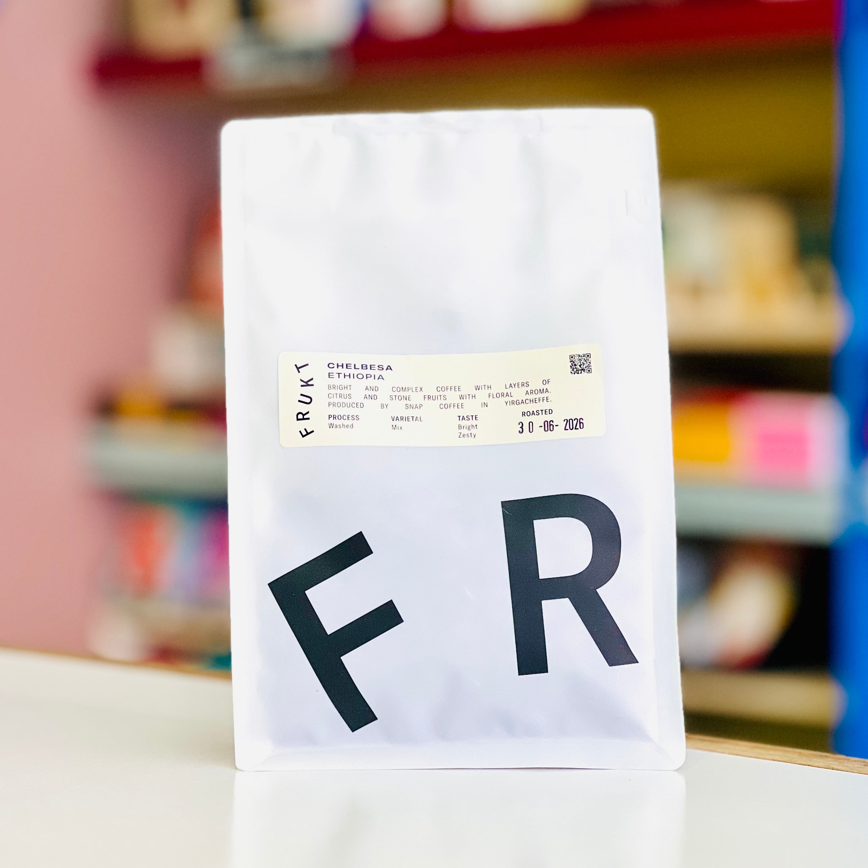 A white coffee bag with large black letters 'F R' on the front and a small product label near the top, standing on a white surface with a blurred indoor shop background.