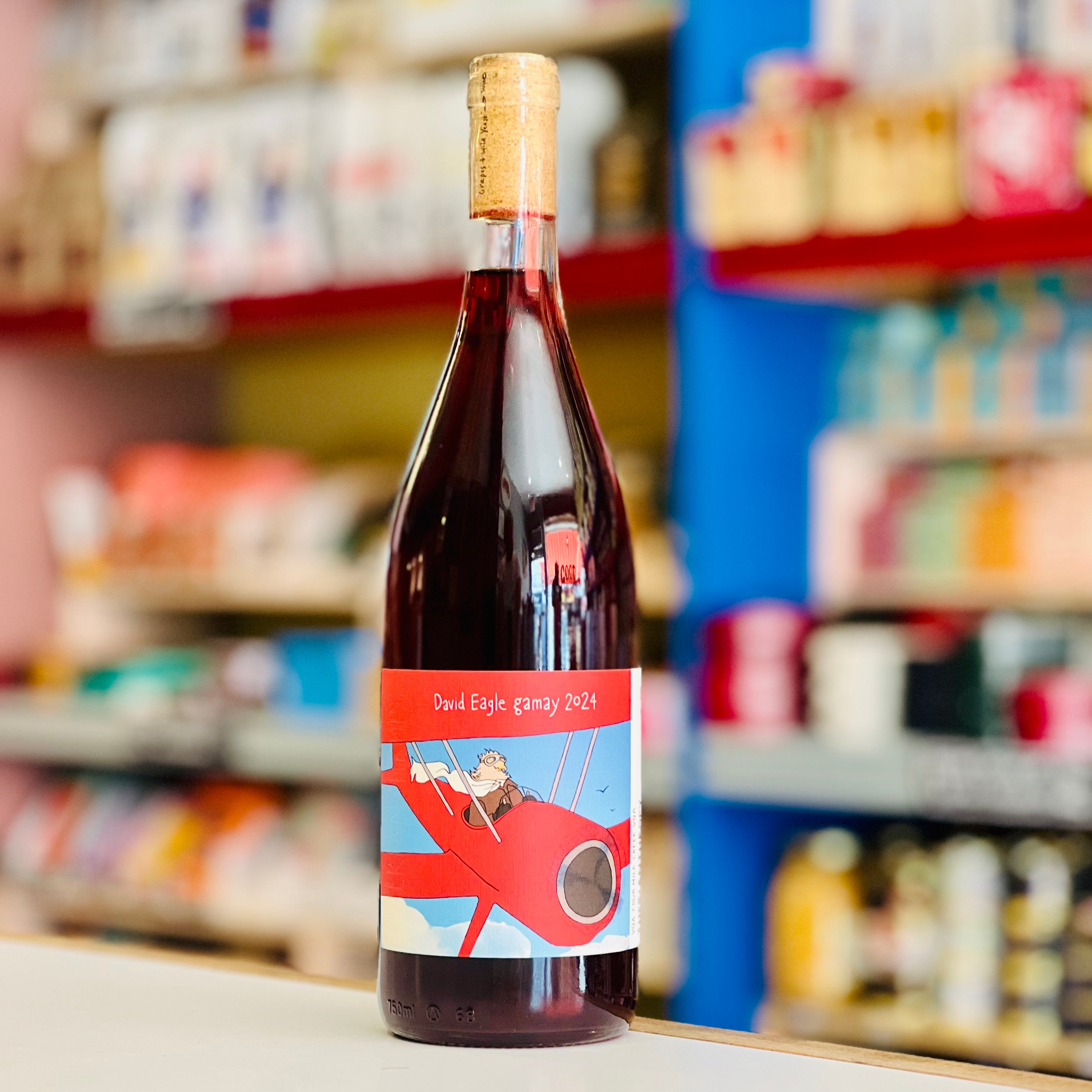 A bottle of red wine with a custom illustrated label featuring a red airplane and the text 'David Eagle Gamay 2024', set against a blurred retail store background.