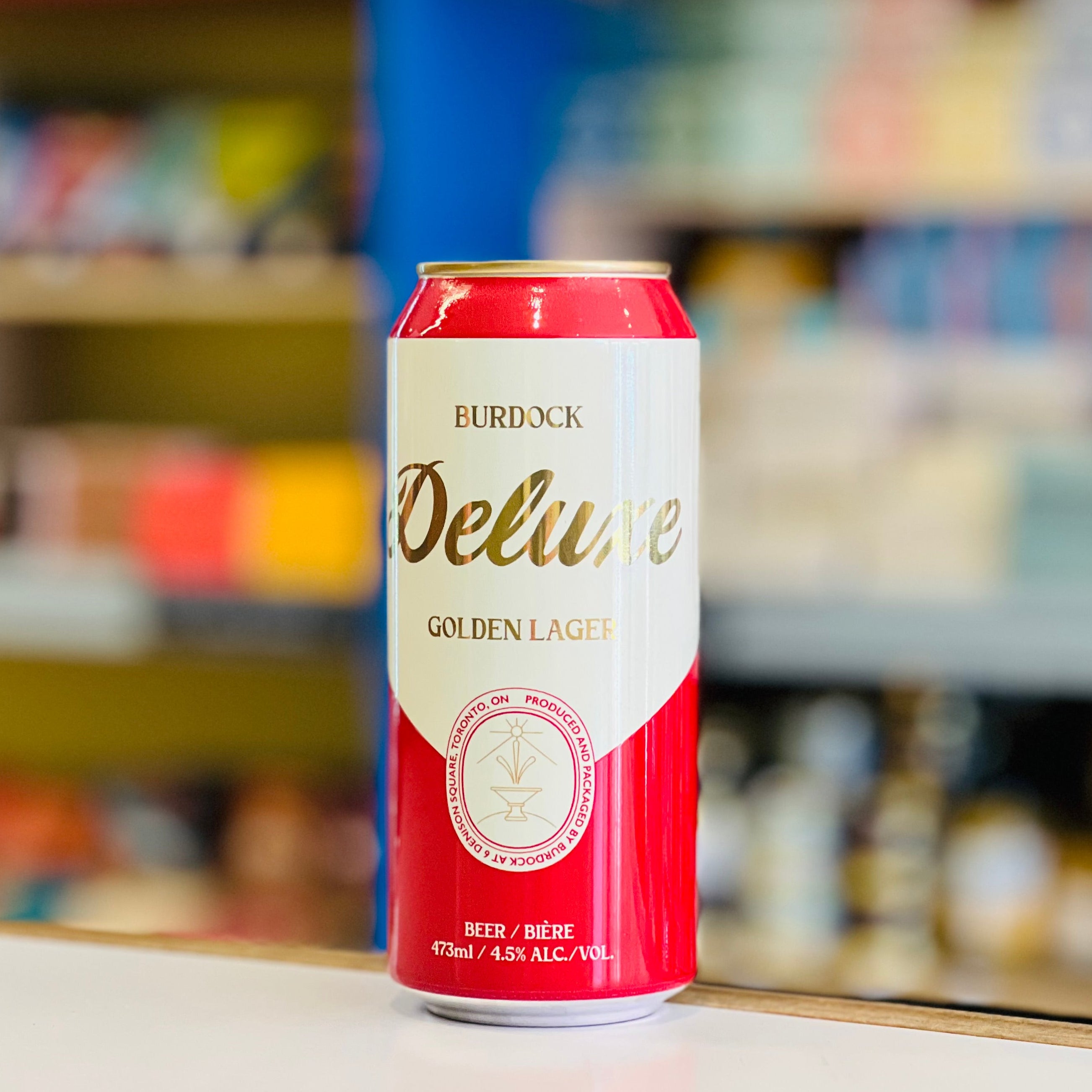 Red and white beer can labeled 'Burdock Deluxe Golden Lager' on a shelf with blurred store shelves in the background.