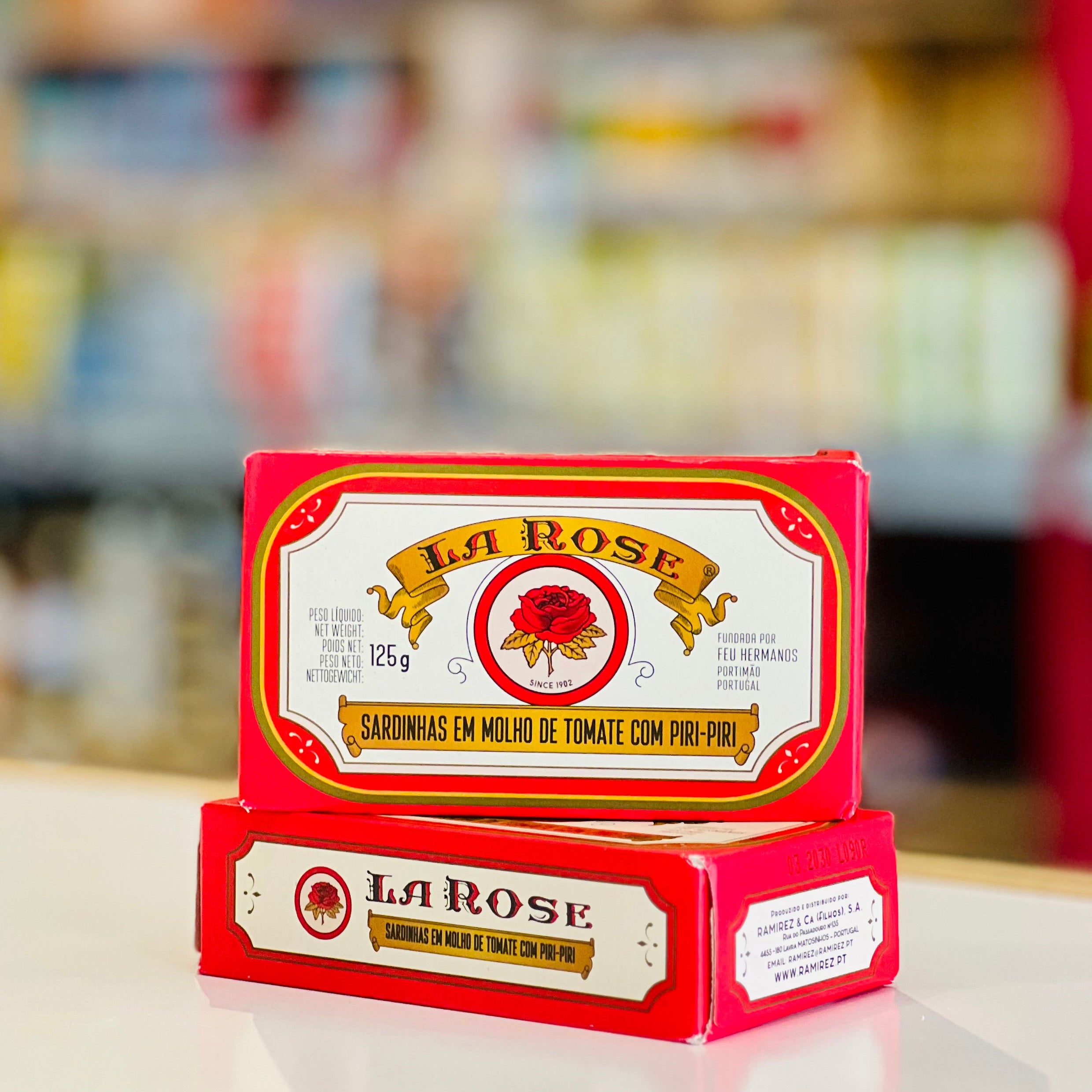 Two red boxes of La Rose Sardinhas em Molho de Tomate com Piro-Piro stacked on a white surface with a blurred store background.