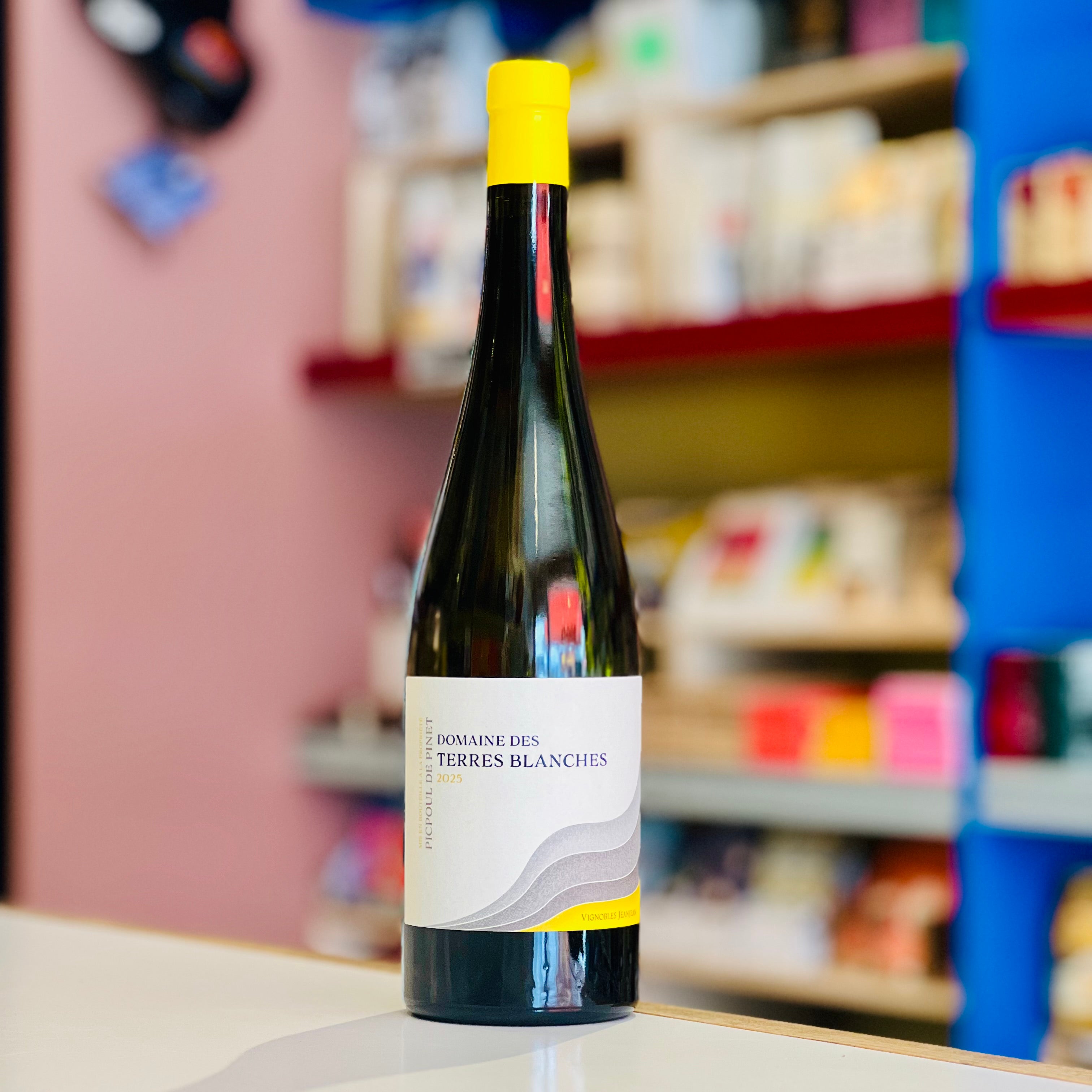 A bottle of wine from Domaine des Terres Blanches stands on a white surface in a shop setting with blurred shelves and colorful walls in the background.