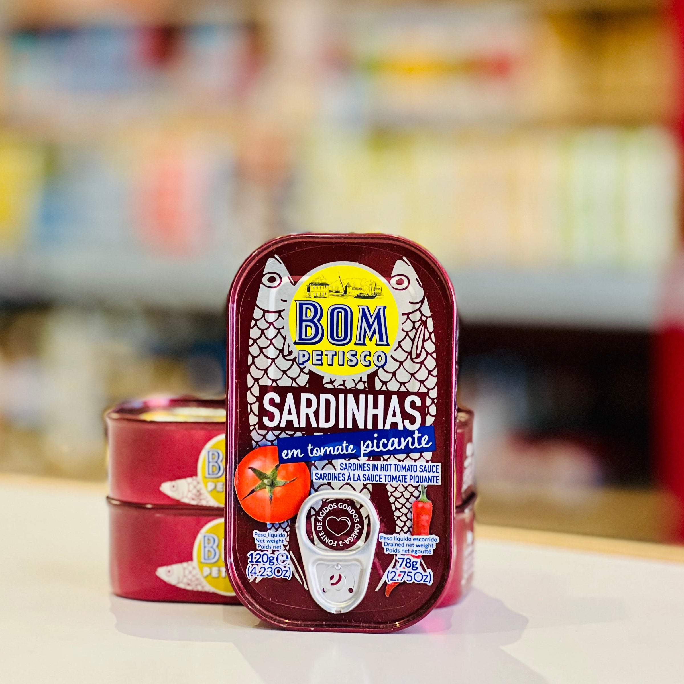 A can of BOM Presão Sardinhas em Tomate Picante canned fish stands on a white surface with two more cans stacked behind it. The background shows a blurred store or kitchen environment with shelves and colorful items.