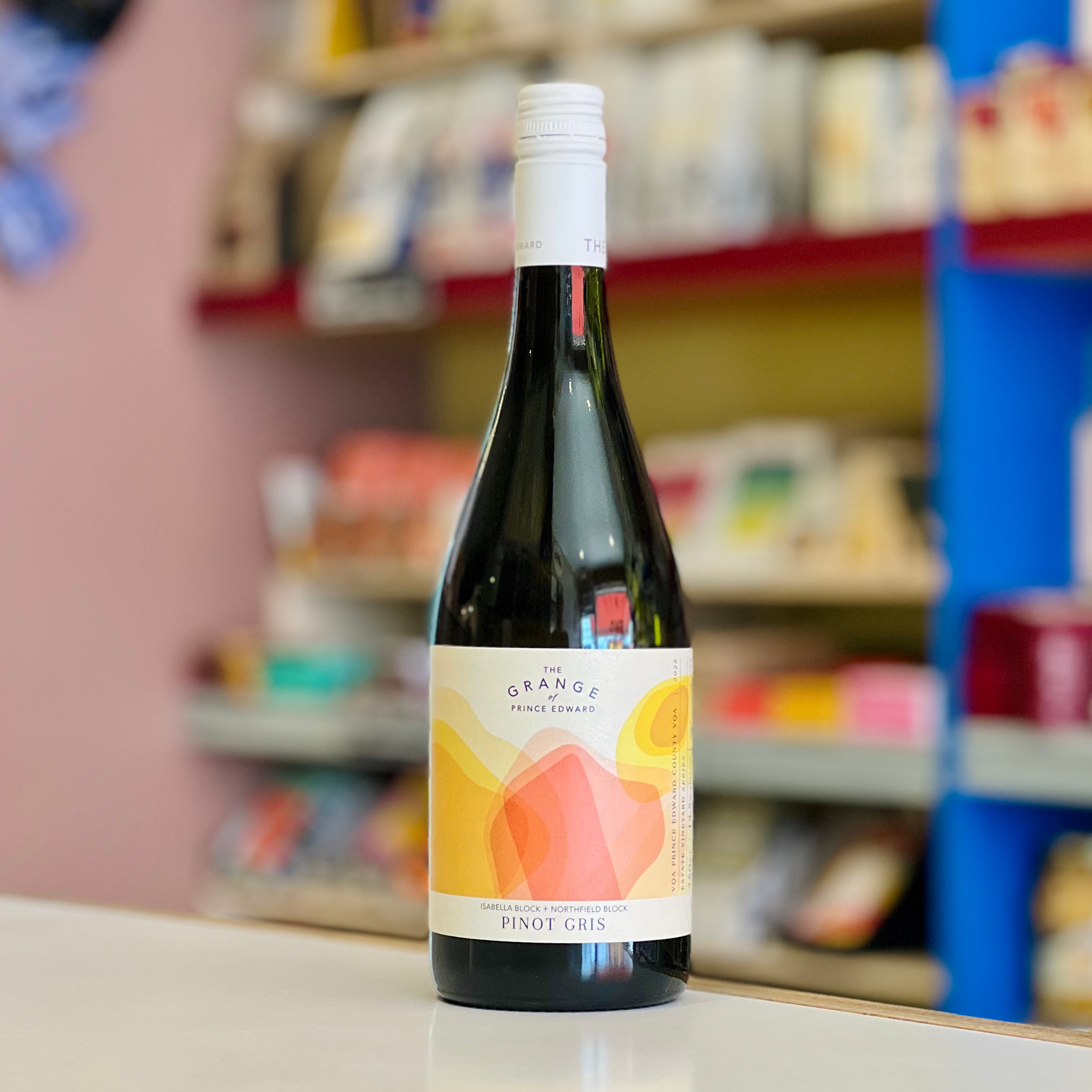 A bottle of Pinot Gris wine from The Orange Farmstead, featuring a white label with orange and yellow geometric graphics, standing on a white counter with a blurred store background.