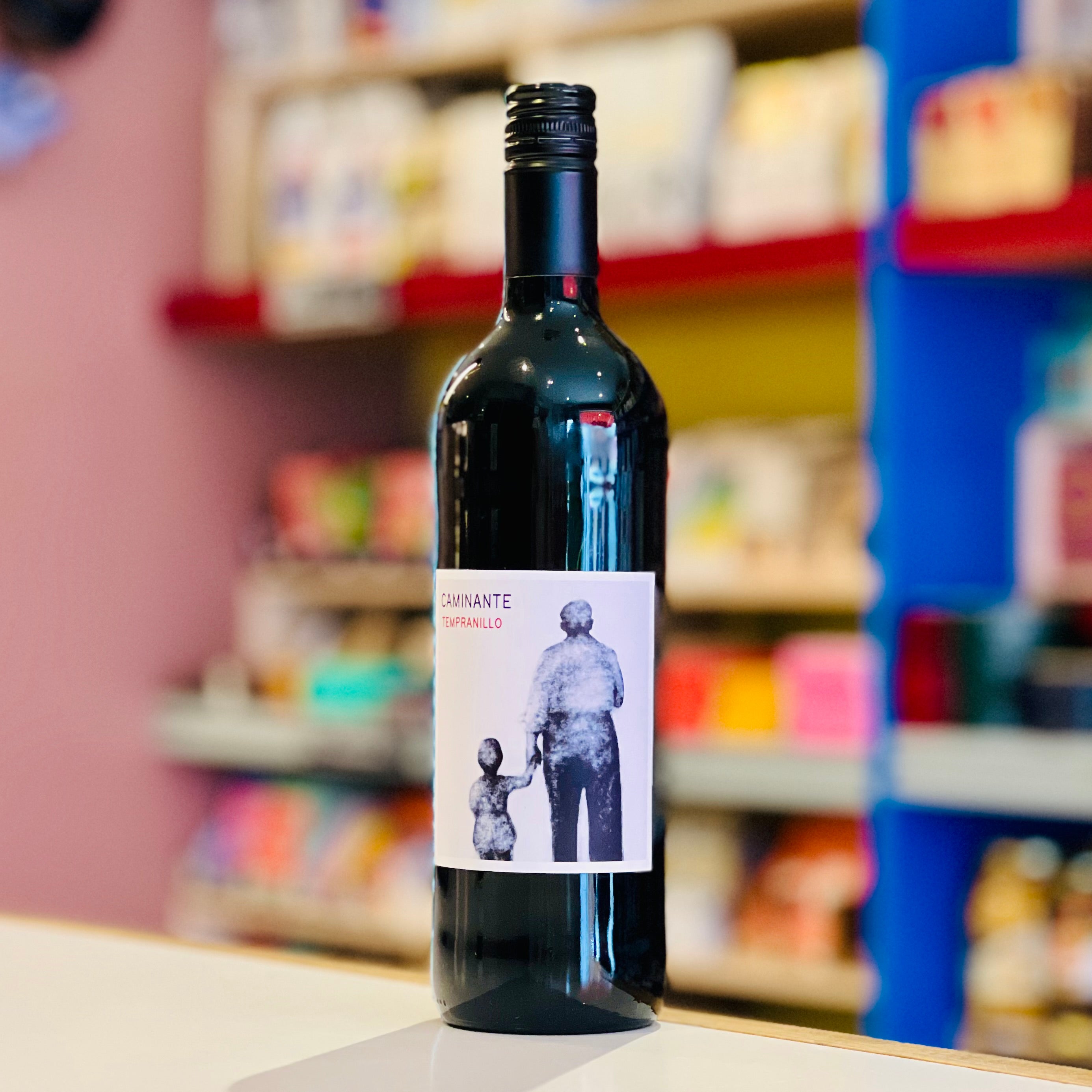A dark glass wine bottle with a black cap and a white label featuring a black-and-white illustration of an adult holding a child's hand, set against a blurred colorful store background.