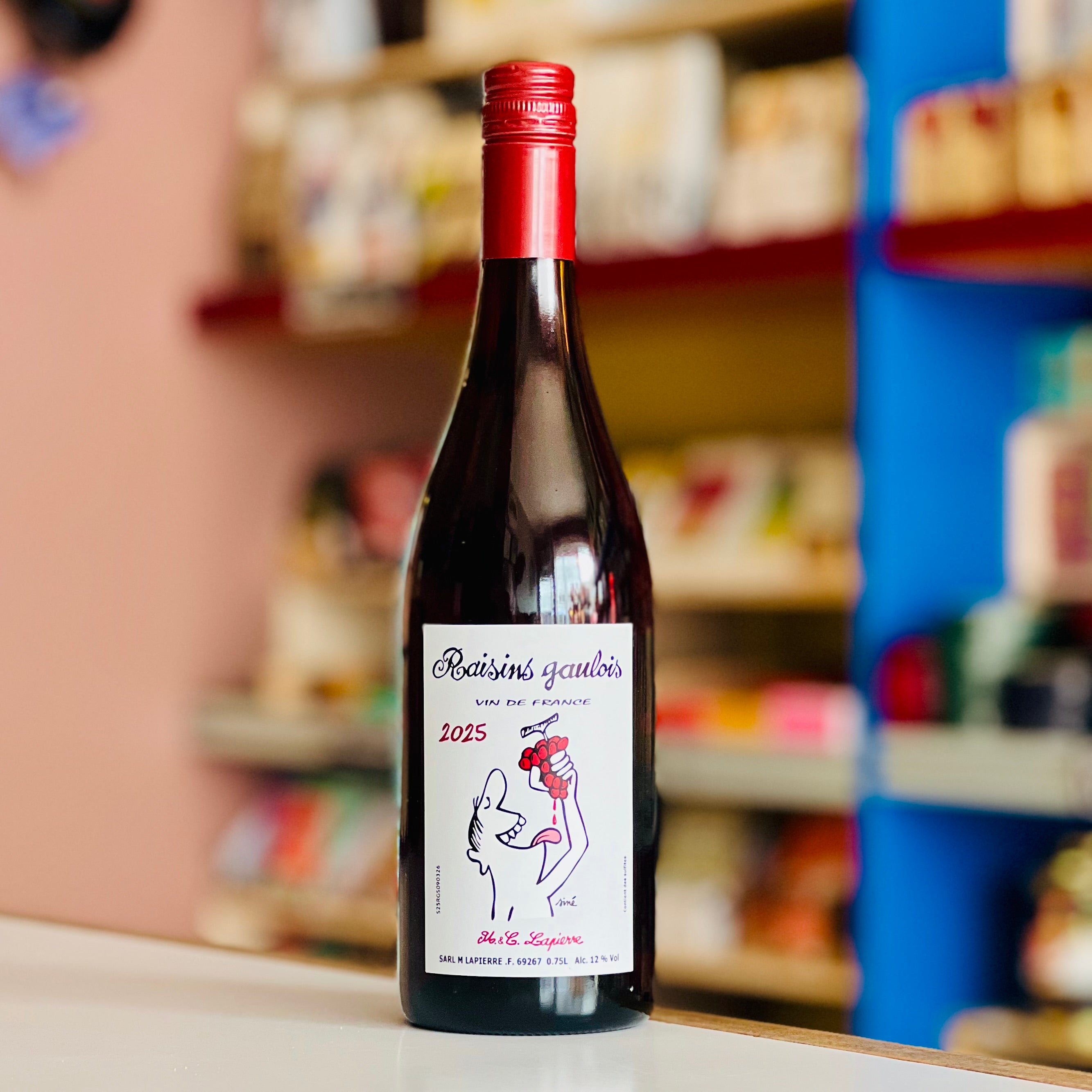 A bottle of Réaumur's Secours wine with a red foil top and a white label featuring a black line drawing of a hand holding a red heart, standing on a white surface with a blurred shop background.