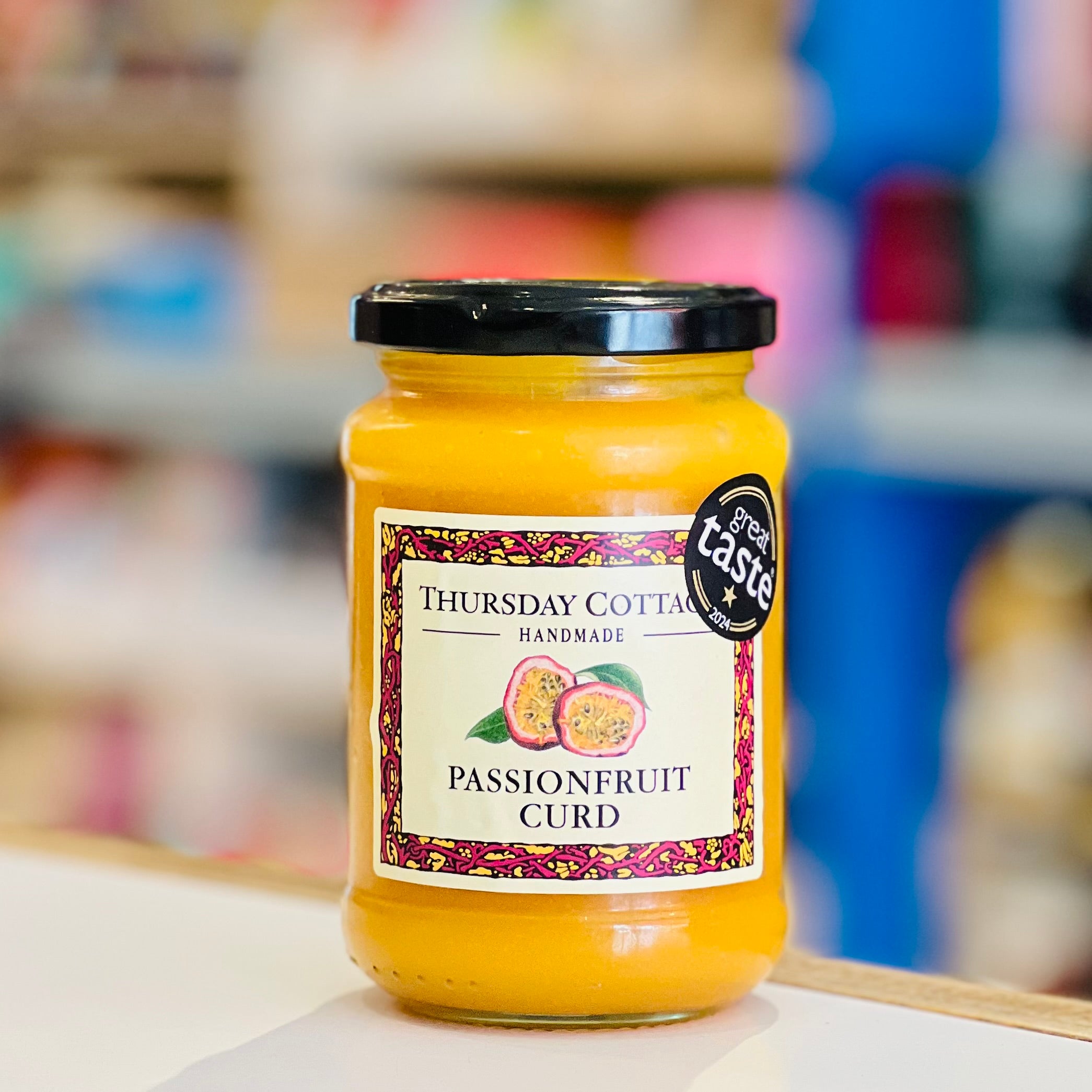 A glass jar of Thursday Cottage Handmade Passionfruit Curd with a black lid and a cream label, set against a blurred background of a shop interior.