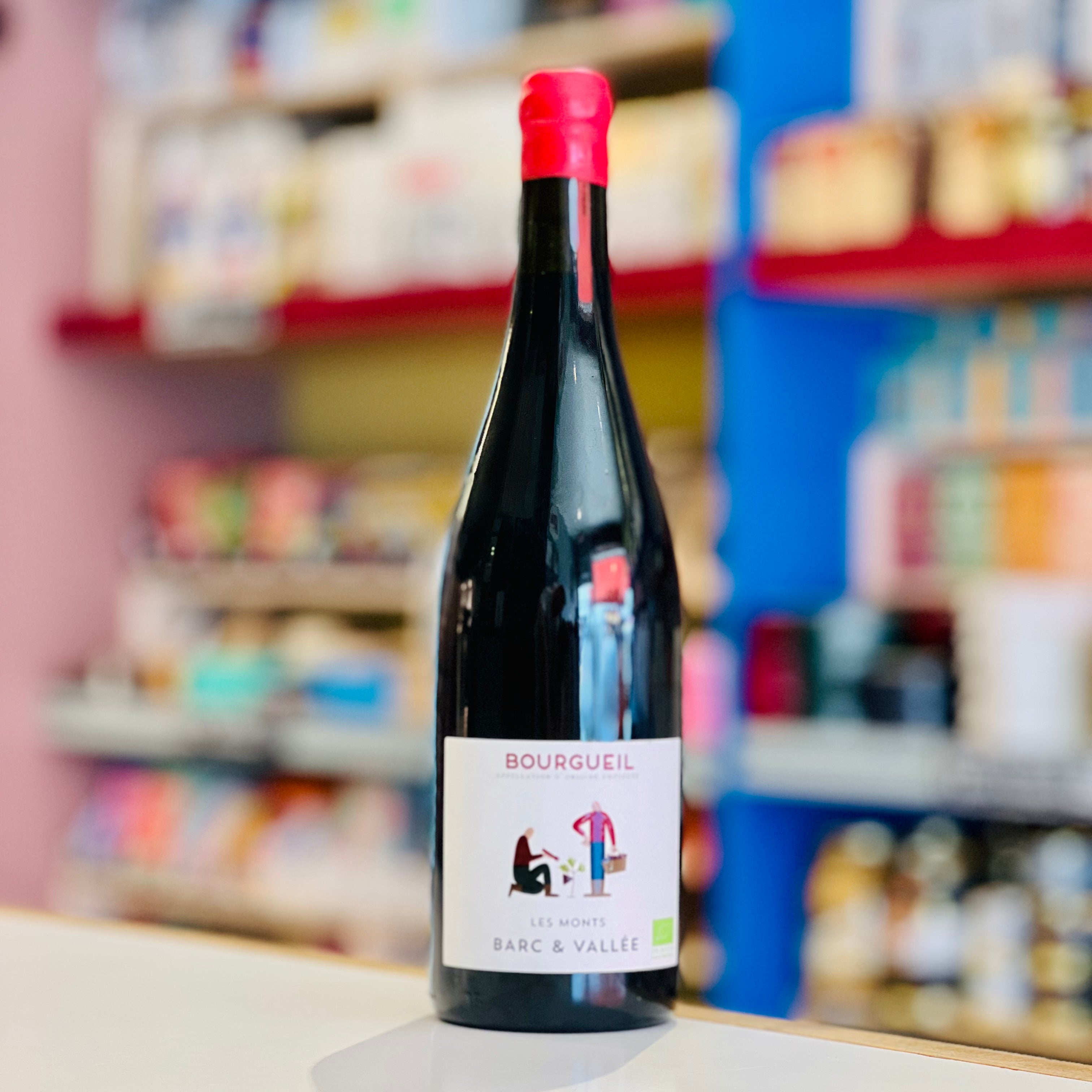A bottle of Bourguel Le Mont Barc & Vallée wine stands on a countertop in a blurred shop setting with colorful shelves in the background.