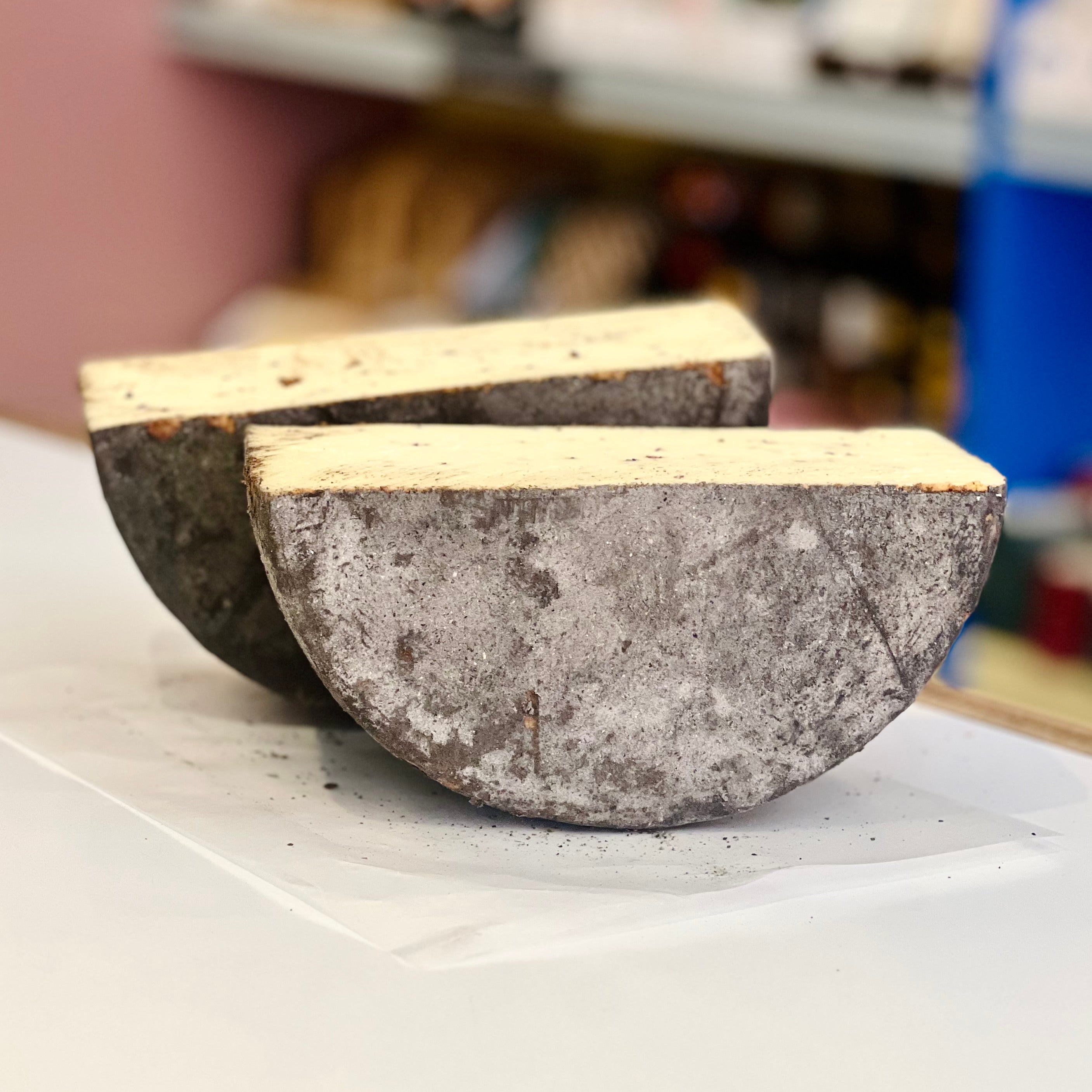 A wheel of cheese with a dark brown rind, cut in half and resting on their side. Blurred background. 