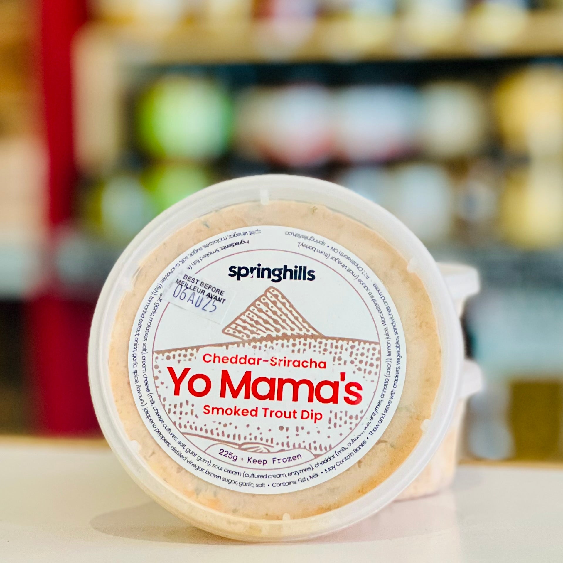 Container of Yo Mama's Smoked Trout Dip with Springhills branding on a blurred store shelf background.