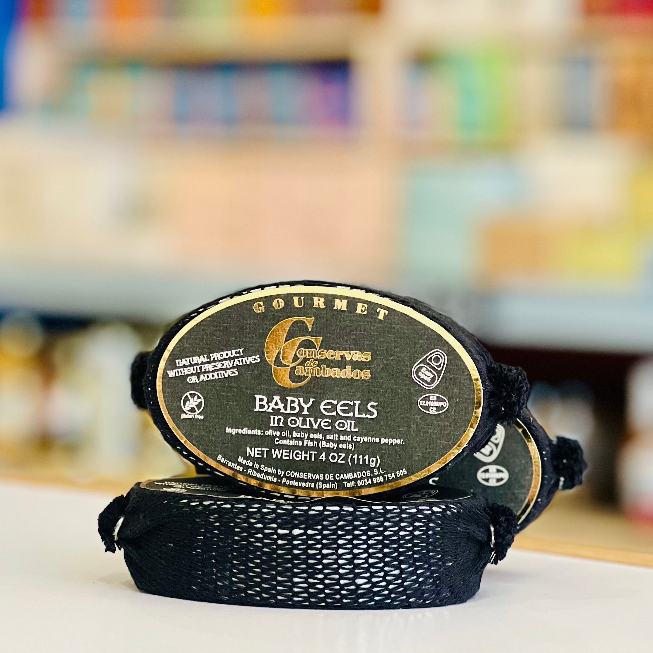 Two black packages of gourmet baby eels in olive oil on a white surface with a blurred store shelf in the background.