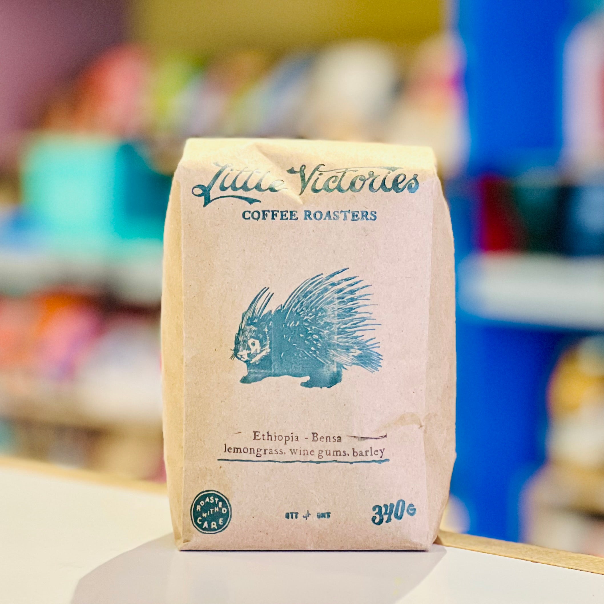 A paper bag of Little Victories Coffee Roasters Ethiopia Bensa, featuring a blue porcupine illustration on a beige background with a blurred shop interior behind it.