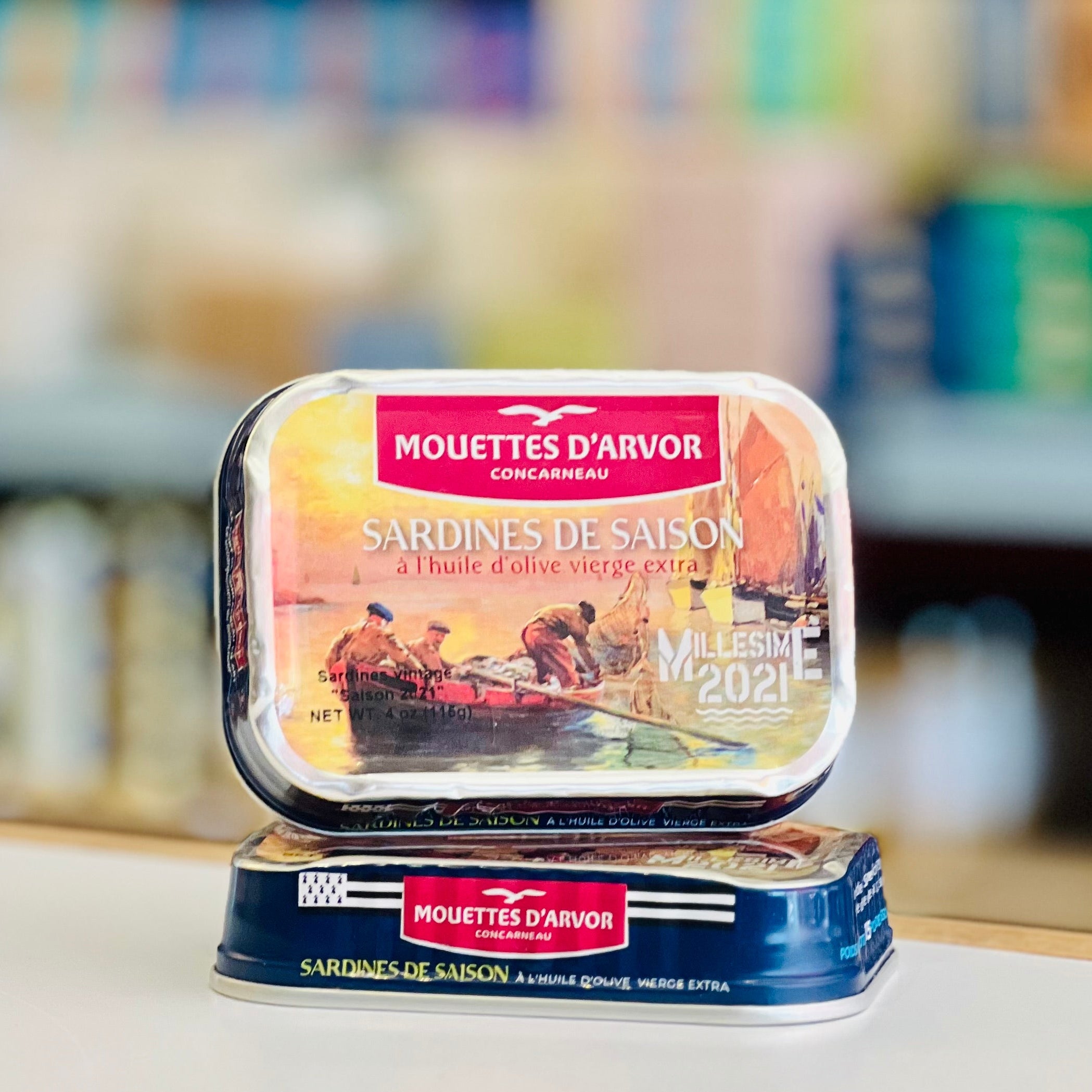 Two tins of Mouettes d'Arvor sardines on a white surface with a blurred store shelf in the background.