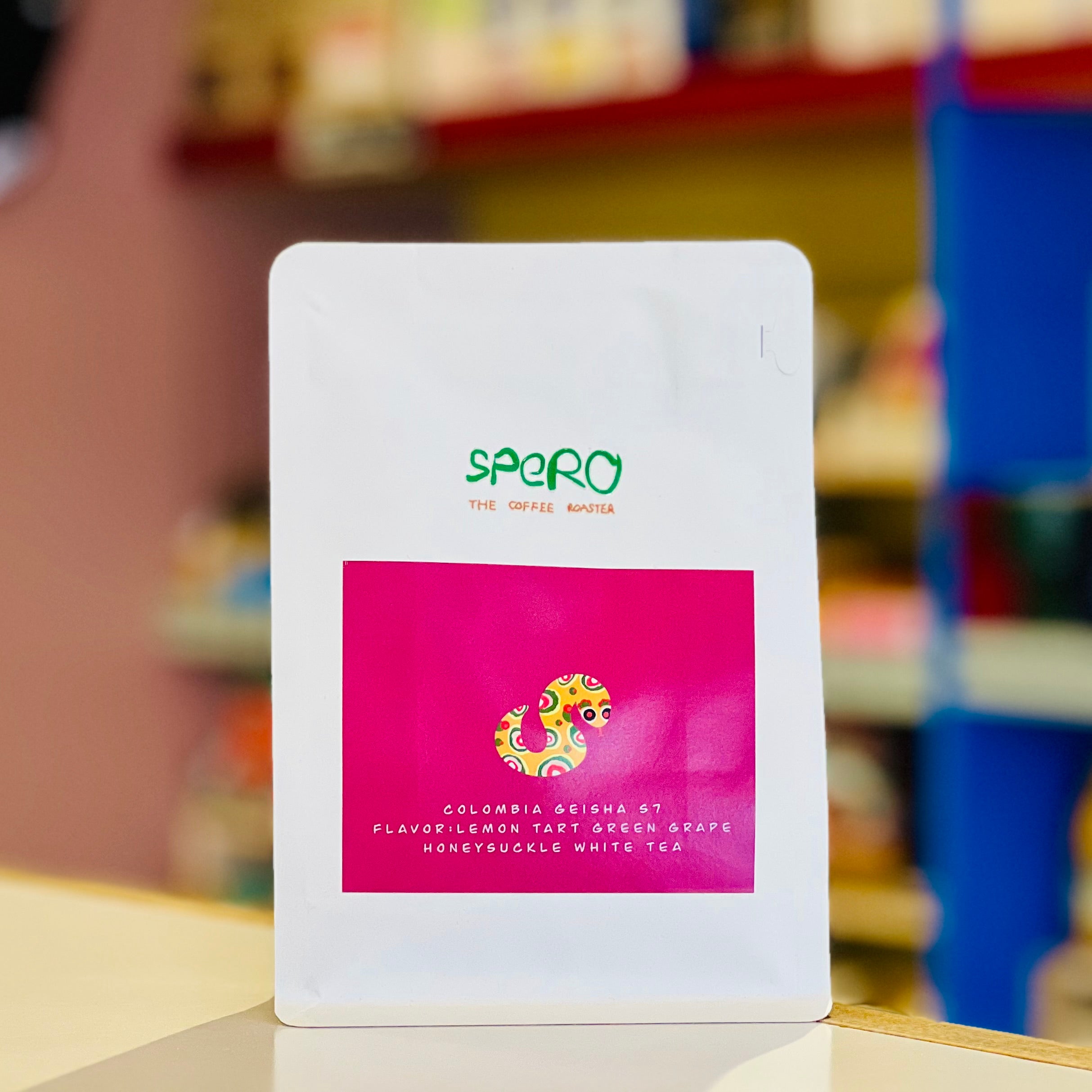 A white stand-up pouch of Spero Coffee Roast tea, featuring a bright pink label with a colorful dragon graphic and text, set against a blurred background of shelves.