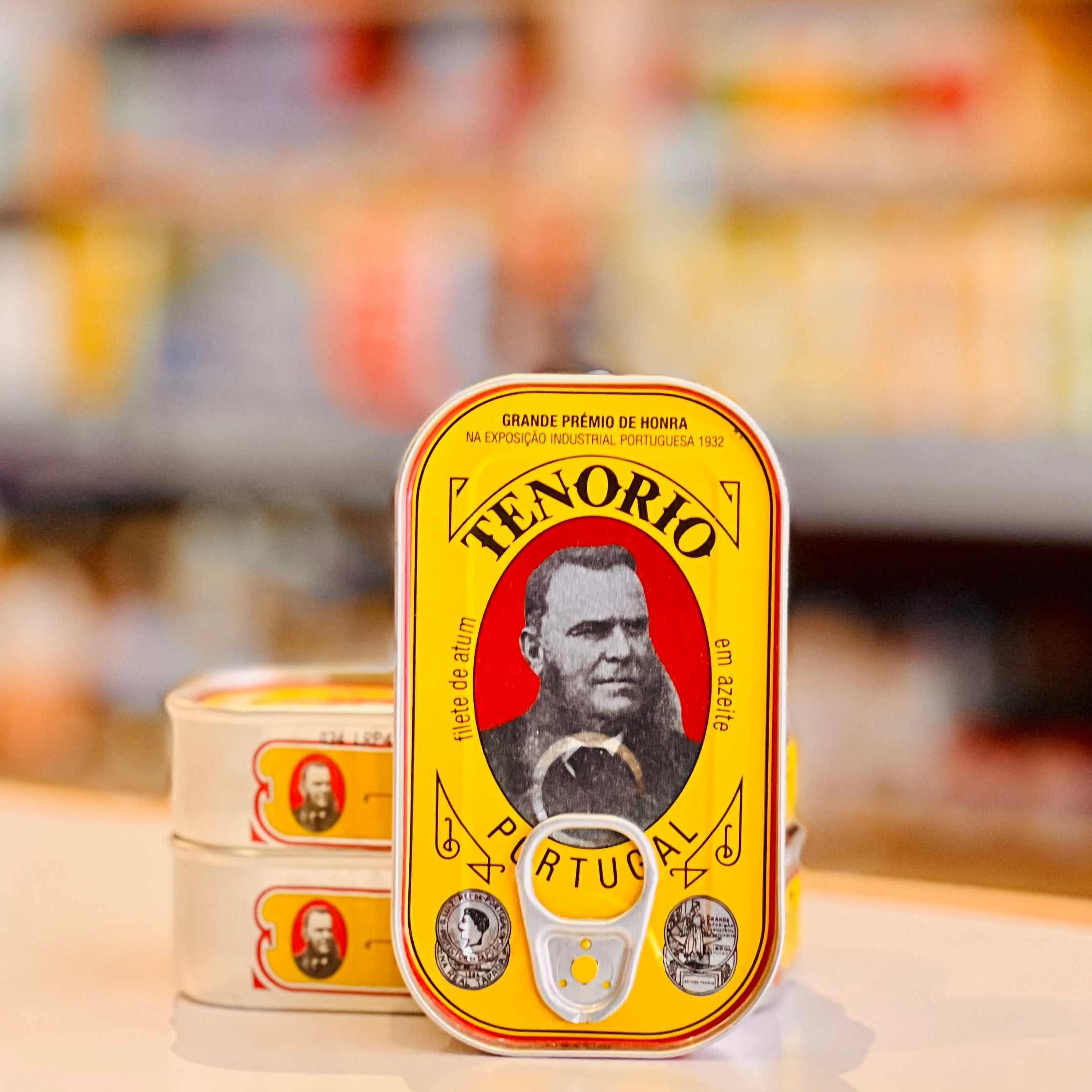 A stack of Tenorio Artucio canned sardines on a light beige countertop, with the top can standing upright in front. The can features a bright yellow label with a portrait of a bearded man and the brand name 'TENORIO' in bold black letters.