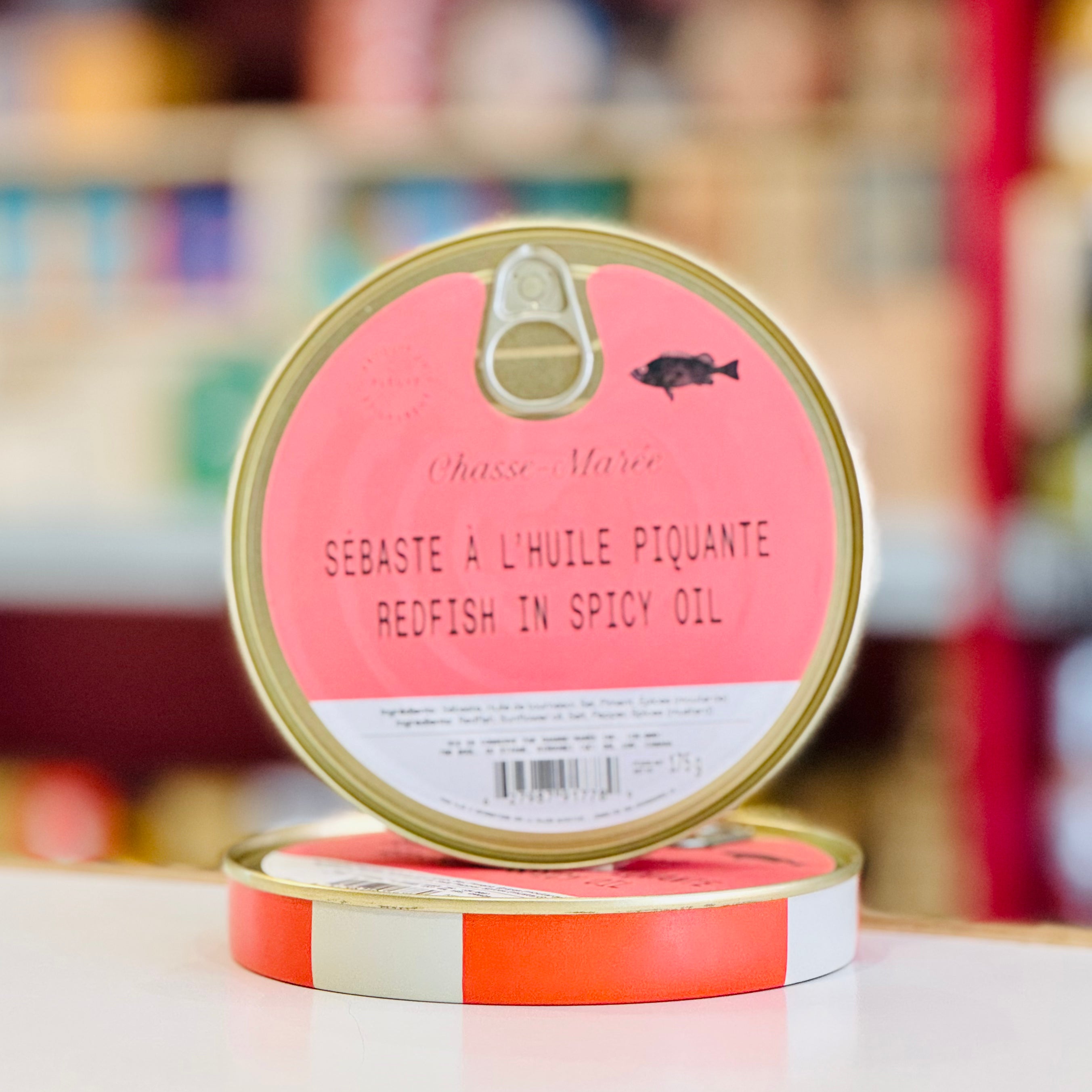Two tins of 'Redfish in Spicy Oil' with a pink label on a blurred background