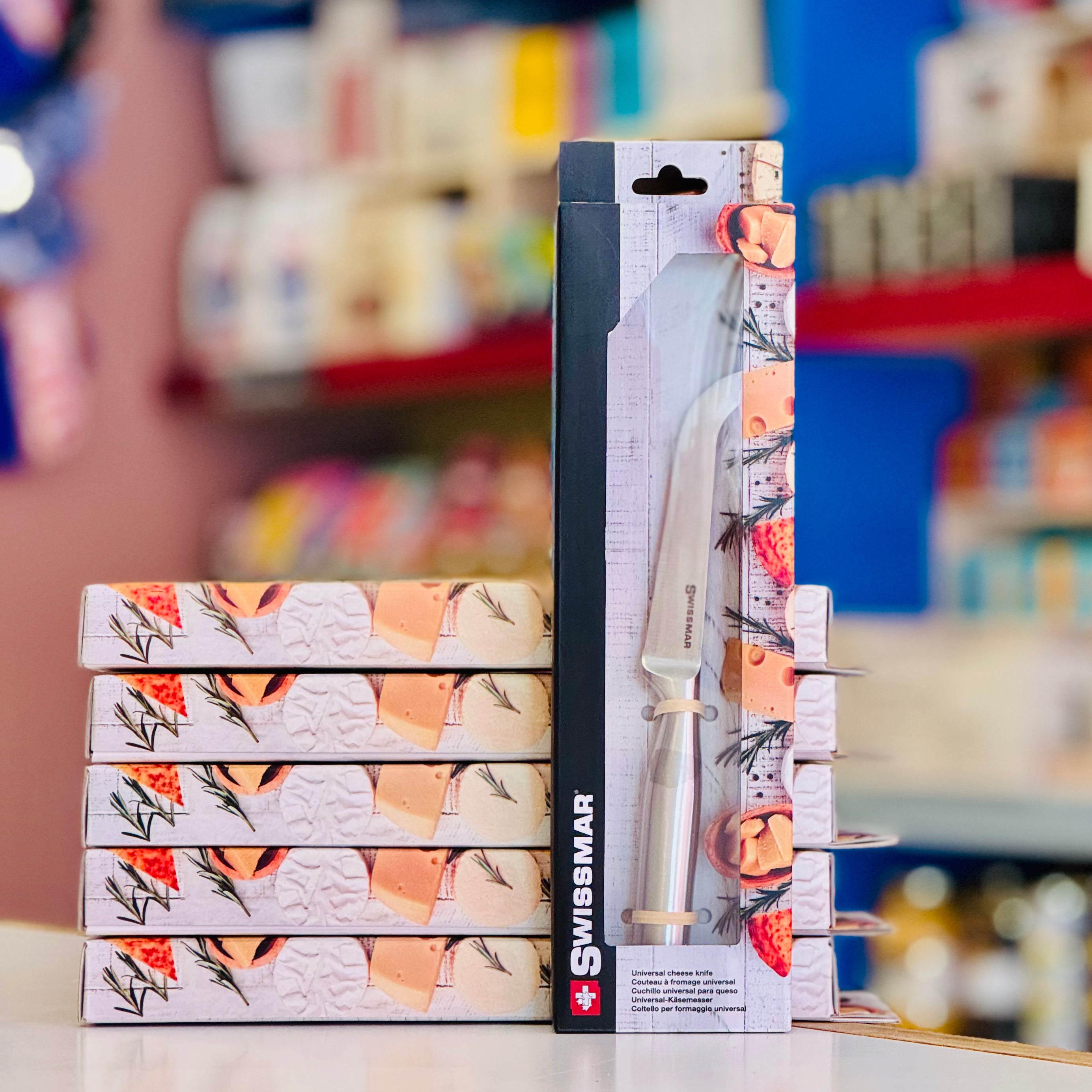 Stack of Swissmar branded knife boxes with a knife on a blurred store shelf background