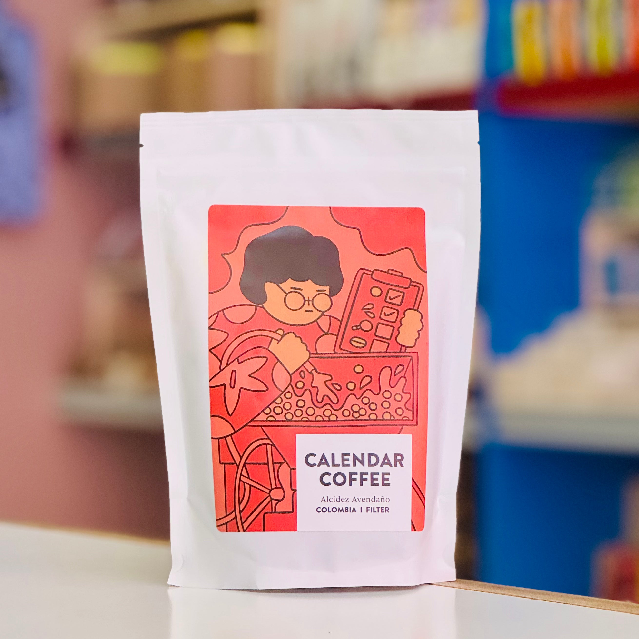 Coffee package with a colorful label featuring an illustration of a person with a calendar, set against a blurred background.
