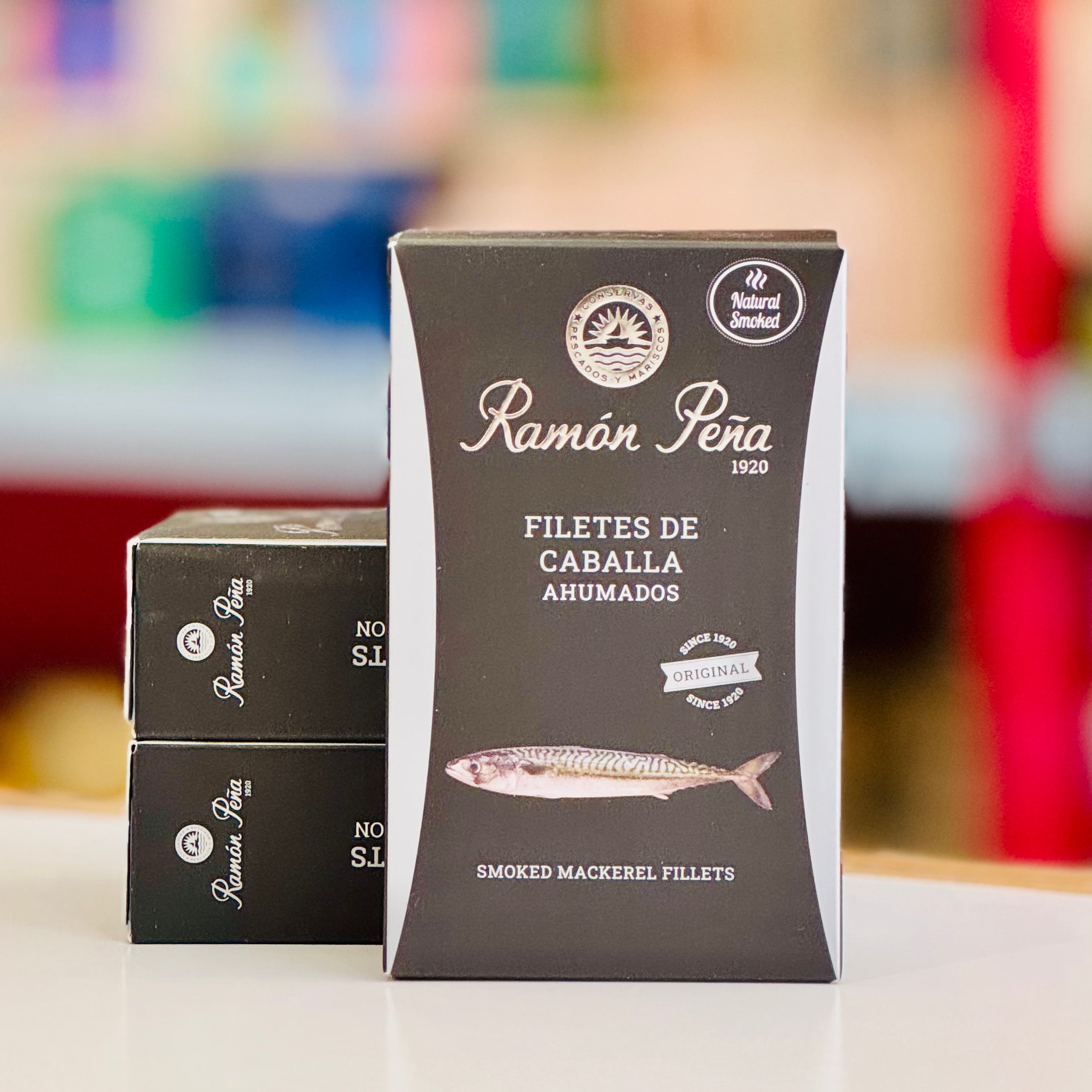 Packaging of Ramón Péra smoked mackerel fillets with a blurred colorful background