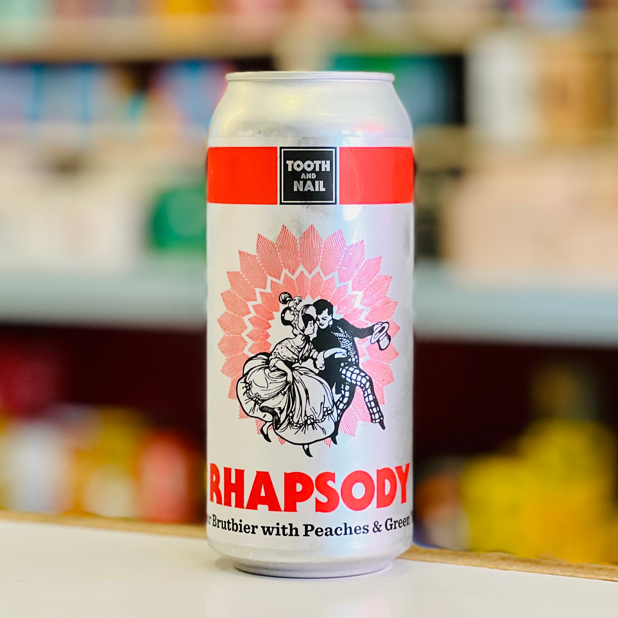 Cans of 'Rhapsody' beer with a colorful label featuring dancing figures on a blurred store shelf background.