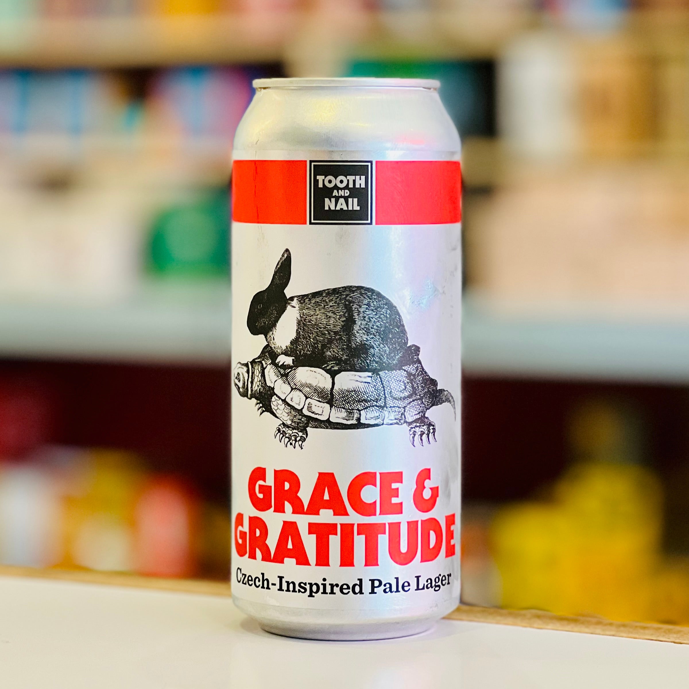 Can of 'Grace & Gratitude' Czech-Inspired Pale Lager with a tortoise and rabbit design on a blurred store shelf background.