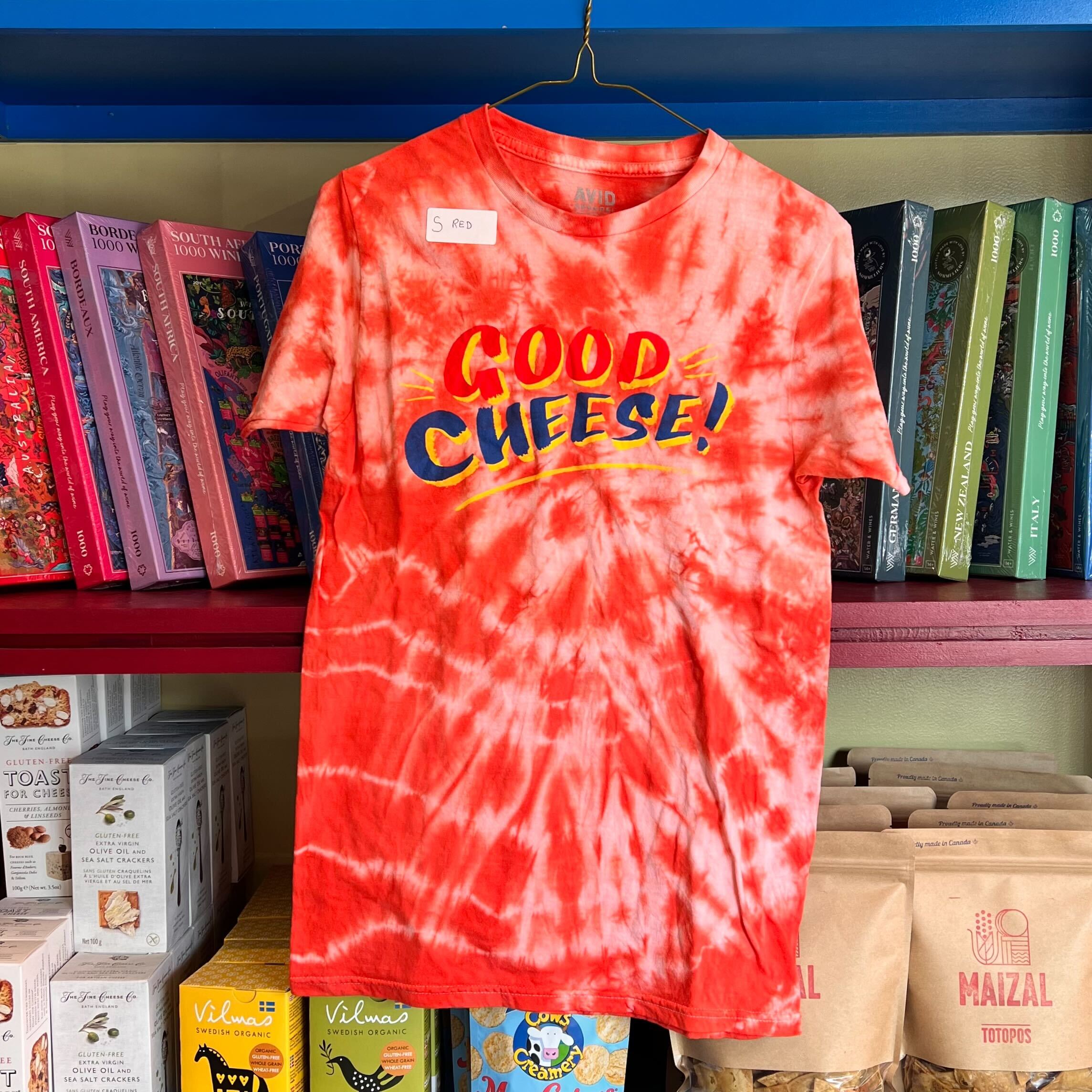 Red and white tie-dye t-shirt with 'Good Cheese!' text on a hanger against a bookshelf background.