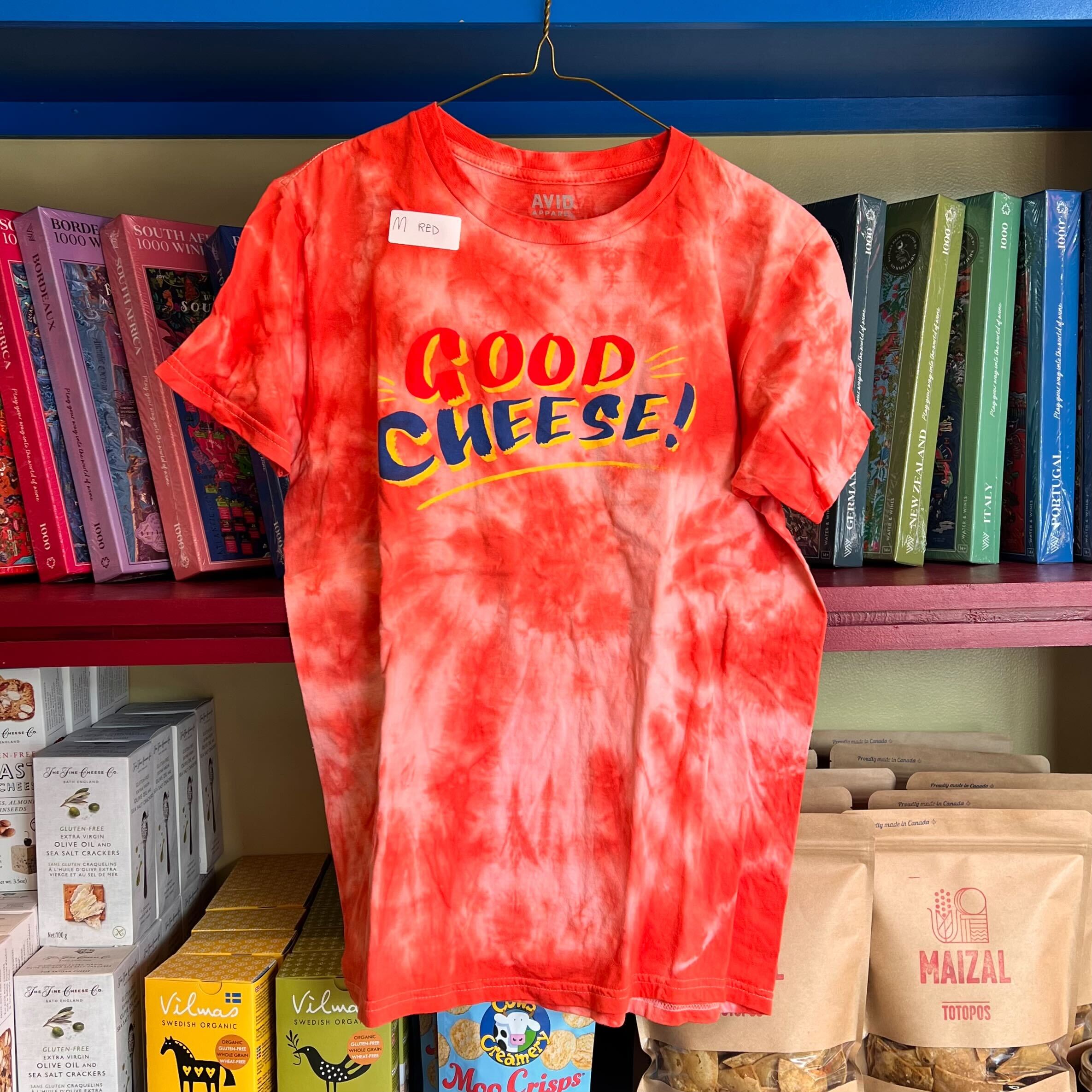 Red tie-dye t-shirt with 'GOOD CHEESE!' text on a hanger against a bookshelf background.