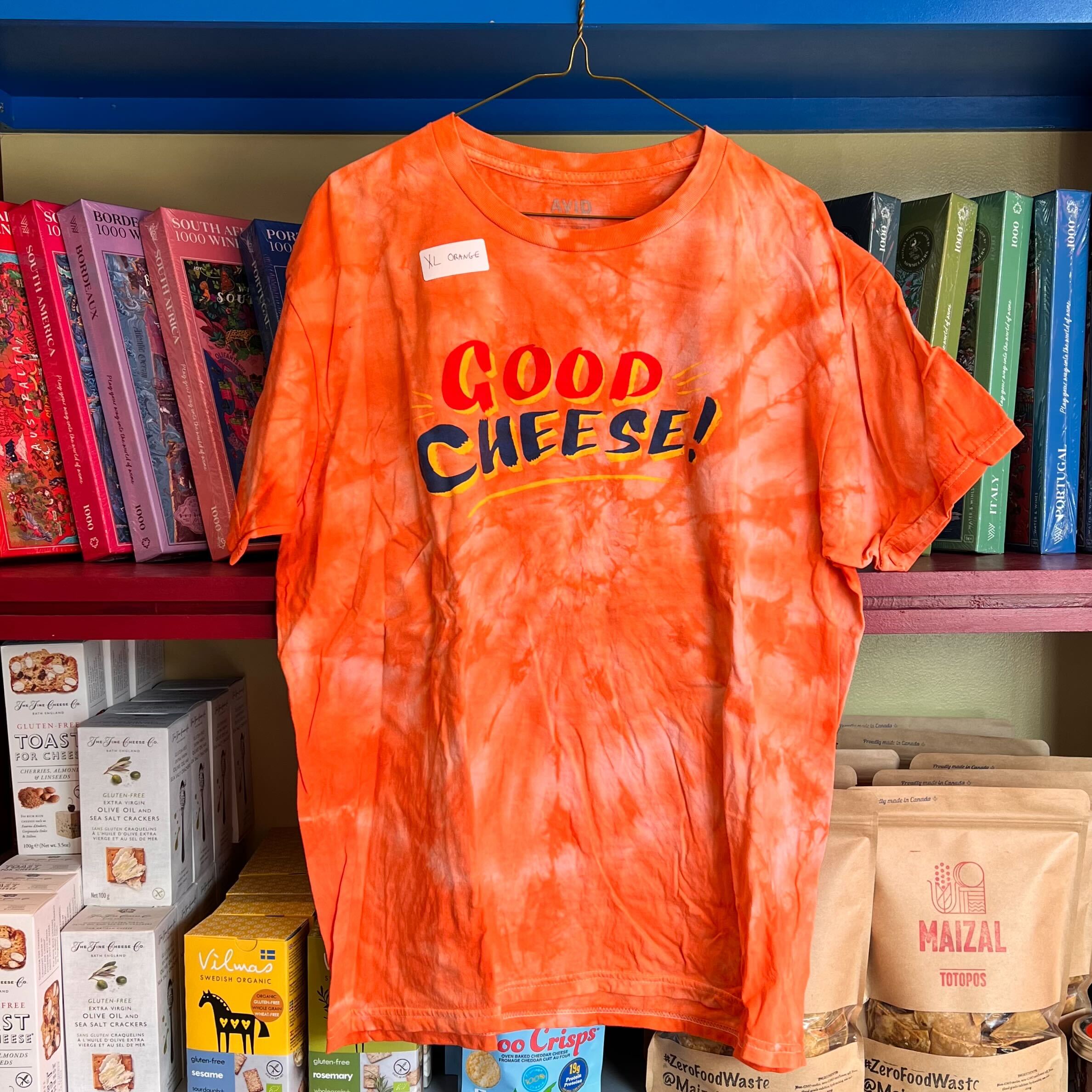 Orange tie-dye t-shirt with 'Good Cheese!' text on a hanger against a shelf background.